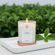 Lit Green Tea Scented Soy Candle in a frosted glass jar with rose gold base, set against a lush green tea leaf backdrop with a fresh sprig alongside