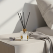 Lavender LUMI Reed Diffuser by Lumi Candles PH on a linen bedroom nightstand beside a dried lavender sprig and a folded grey linen cloth in soft morning light