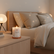 Lit Sandalwood Dreams woody scented candle glowing on a bedroom nightstand beside a cozy bed